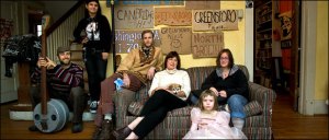 Liz Seymour, in white, sitting beneath a collection of hitchhiking signs, began an experiment in group living at age 52. Five of her six housemates are pictured; the youngest is Skye Tull, 6.